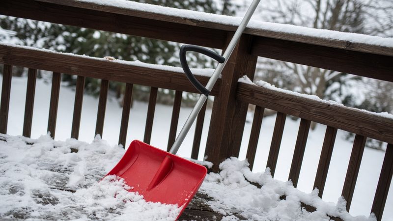 A red snow shovel with a second curved handle leaning on a snowy deck railing, in Rochester, New York